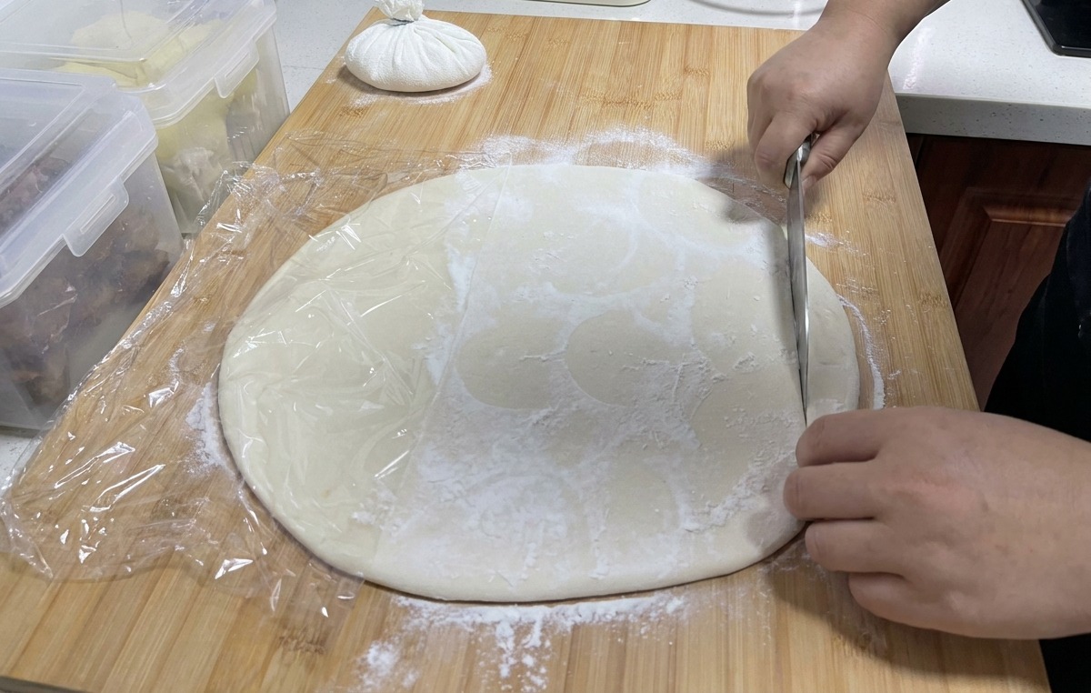 Un grande coltello da cucina che taglia una sfoglia di pasta bianca su un tagliere per creare strisce lunghe.