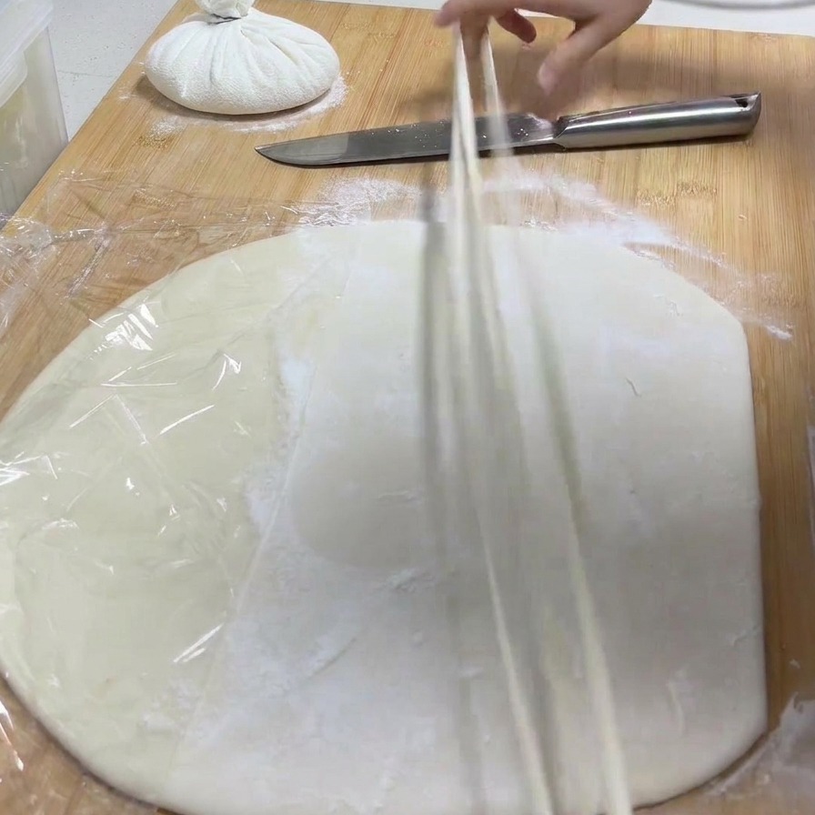 Una mano che solleva una singola striscia di pasta da un tagliere e la allunga trasformandola in un noodle sottile.