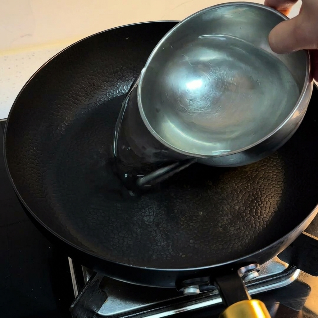 Acqua versata da una ciotolina in una padella antiaderente nera sul fuoco per preparare le uova.