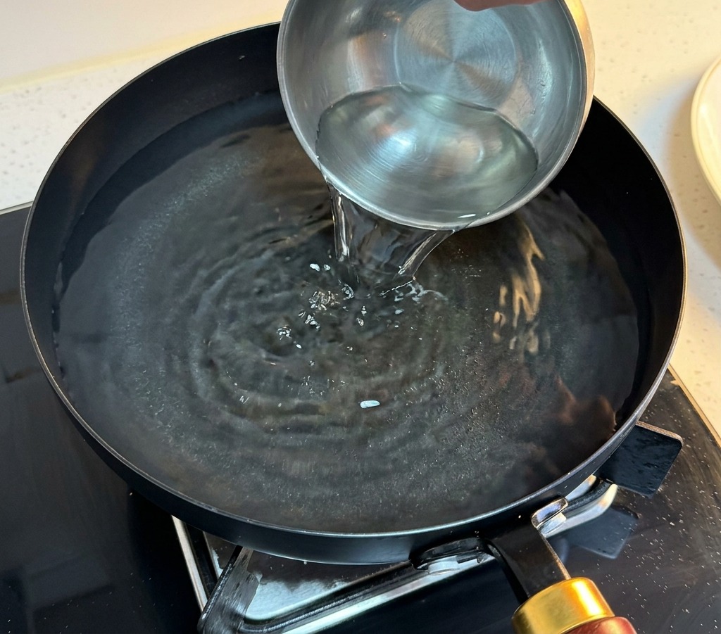 Mano che versa acqua fredda da una piccola ciotola di metallo in una padella di acqua bollente.