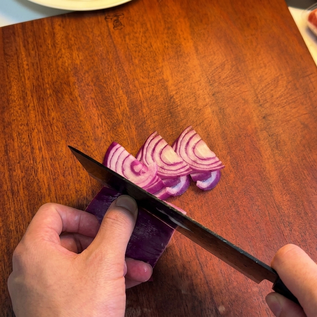 Persona che usa un coltello da chef scuro per affettare una cipolla rossa a spicchi sottili su un tagliere.