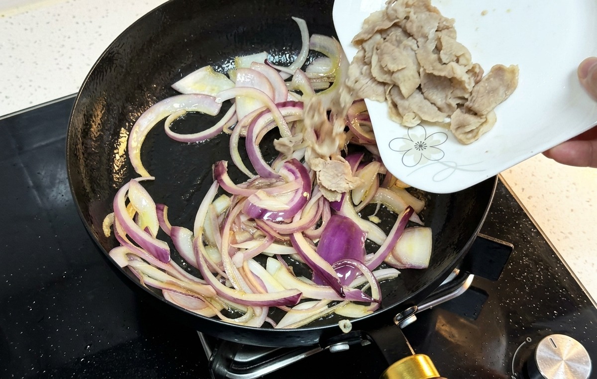 Fette di manzo cotto versate da un piatto bianco in una padella con cipolle rosse soffritte.