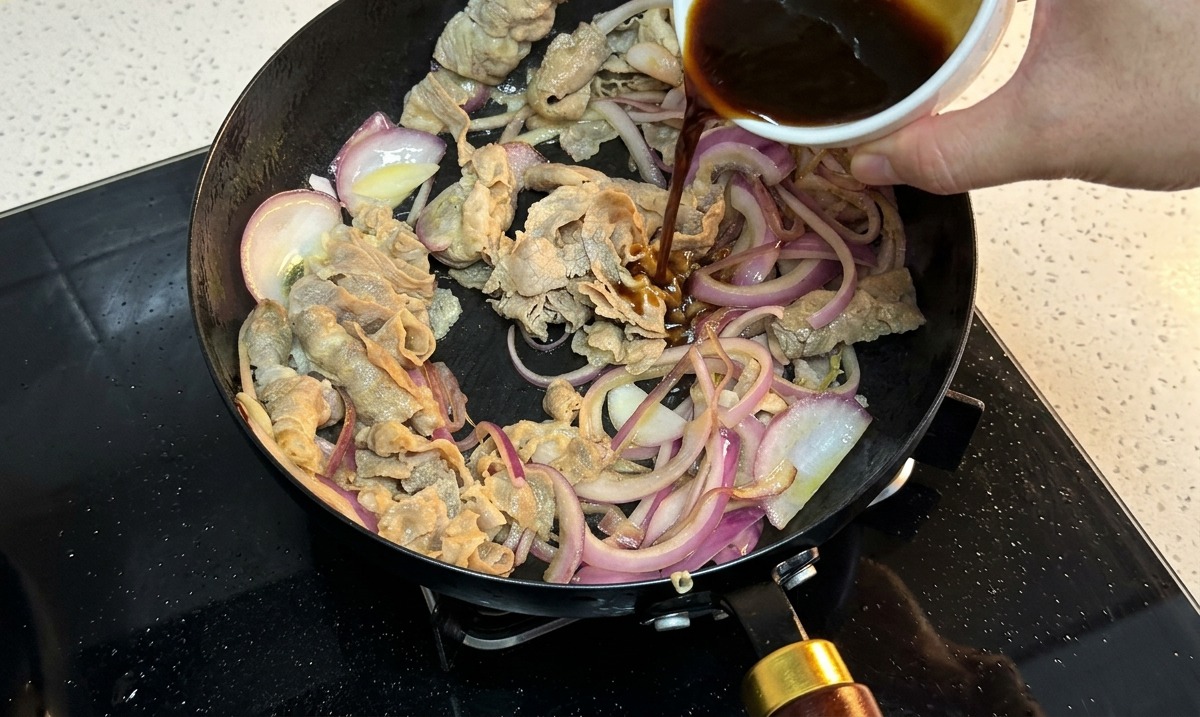 Salsa scura versata da una ciotolina di ceramica in una padella con manzo e cipolle rosse.