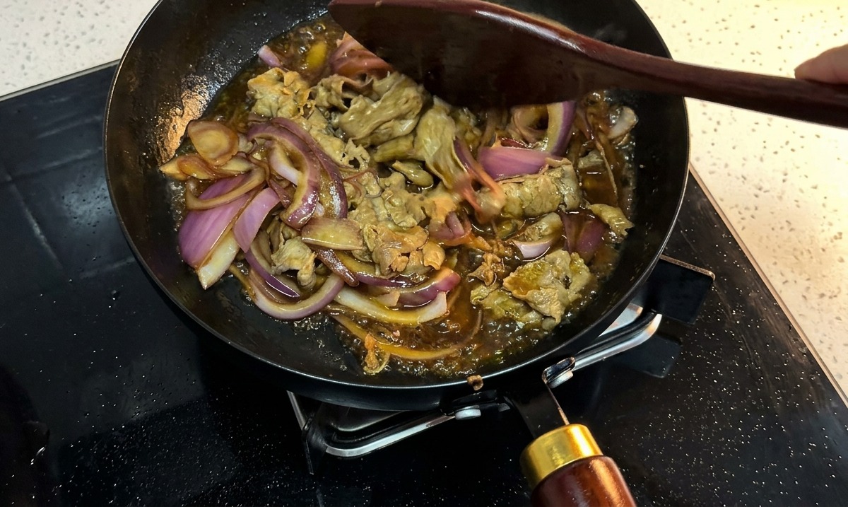 Manzo e cipolle che sobbollono in una salsa scura densa in una padella, mescolati con una spatola di legno.