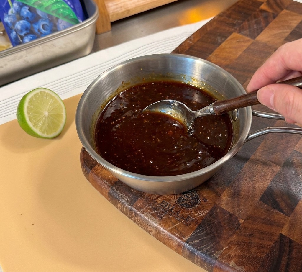 Mano che mescola una salsa teriyaki densa e scura in un pentolino di metallo.