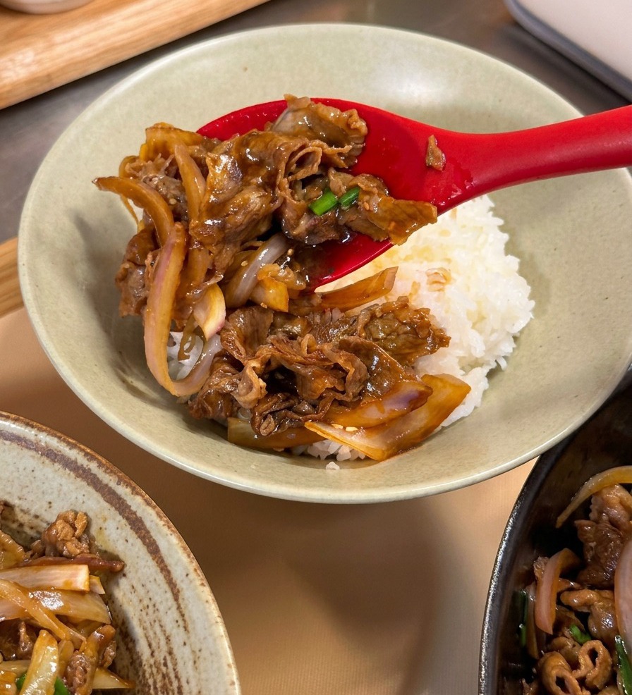 Cucchiaio rosso che adagia il manzo teriyaki su un letto di riso bianco in una ciotola di ceramica.