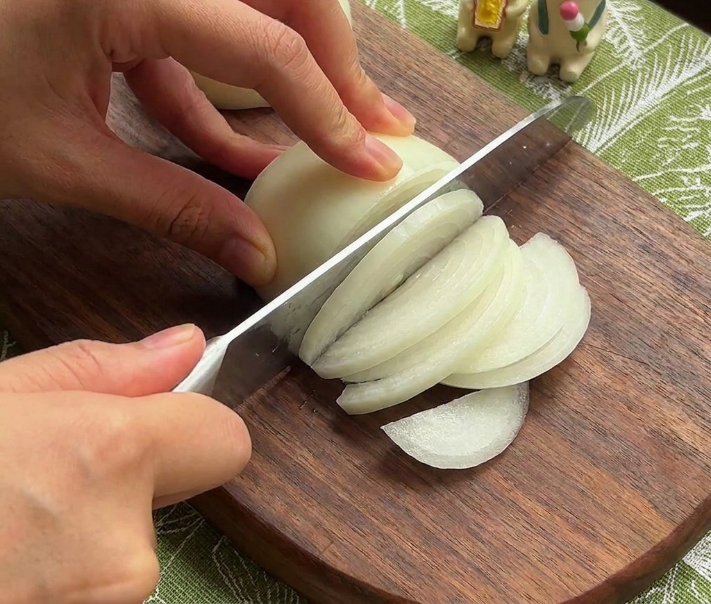 Mani che usano un coltello da chef per affettare una cipolla bianca a strisce sottili su un tagliere di legno scuro.