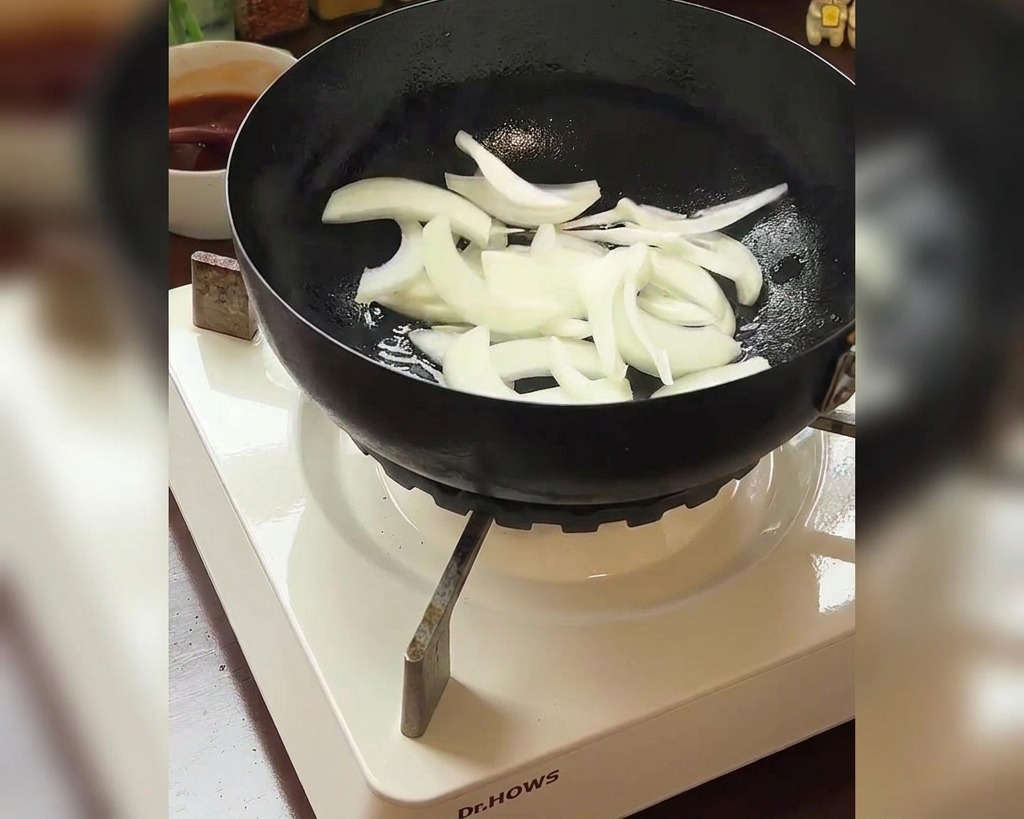 Cipolle affettate aggiunte in padella con una ciotola di kimchi rosso visibile sullo sfondo.