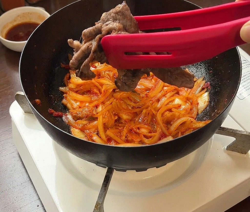 Fettine di manzo rimesse in padella con kimchi e cipolle usando delle pinze rosse.