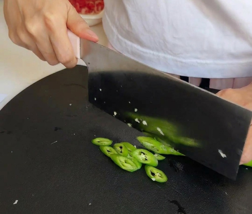 Un coltello da cucina che affetta peperoncini verdi brillanti su un tagliere nero testurizzato.
