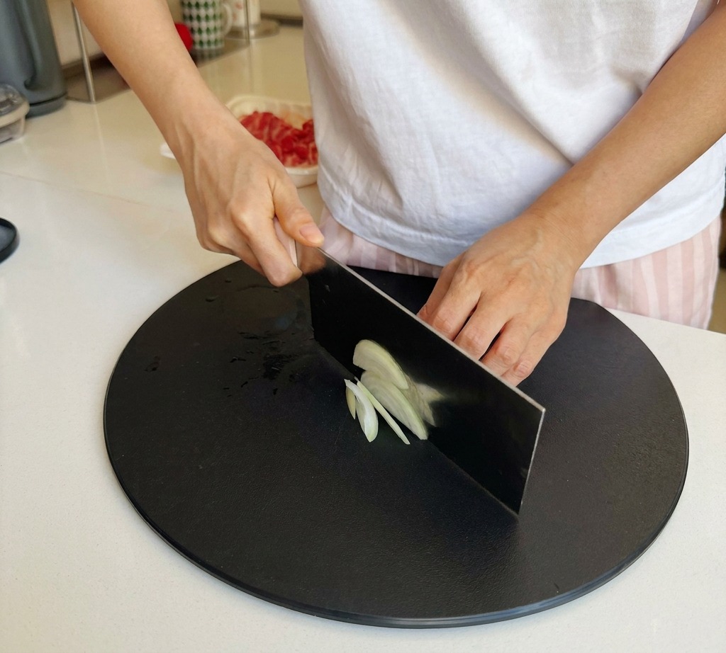 Mani che utilizzano un grosso coltello da cucina per affettare una cipolla bianca in strisce sottili su un tagliere nero.