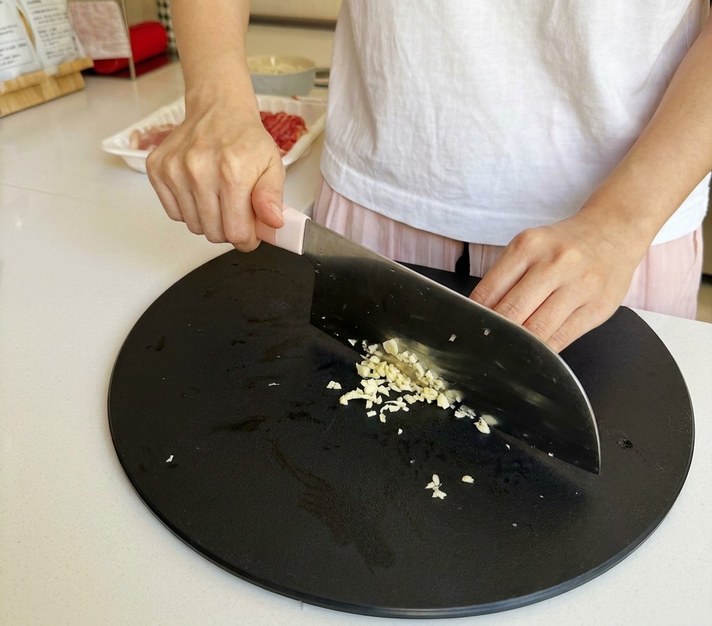 Primo piano di mani che tritano finemente spicchi daglio su un tagliere nero usando un coltello da chef.
