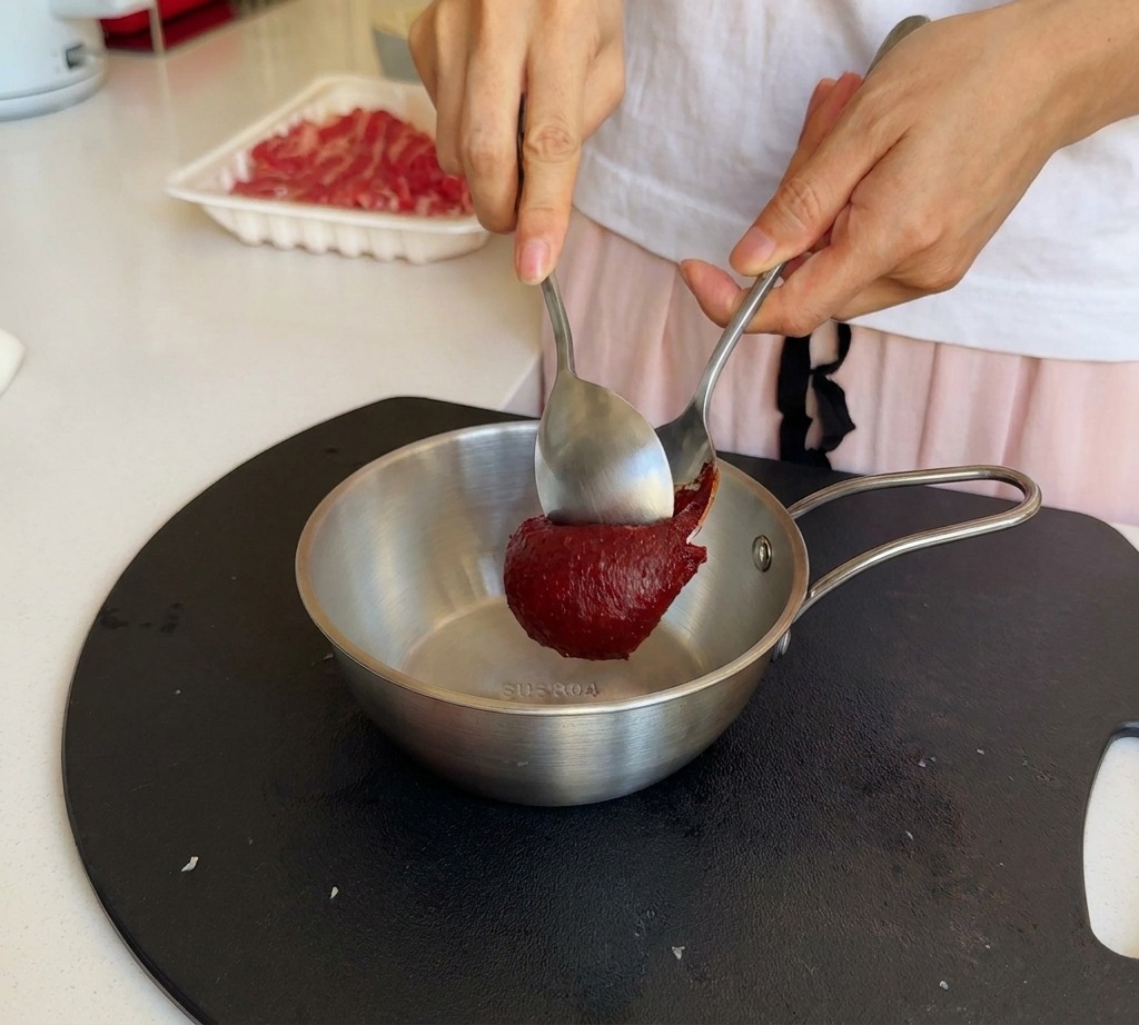 Una persona usa due cucchiai di metallo per aggiungere una generosa cucchiaiata di pasta di peperoncino coreana in una ciotola dacciaio.