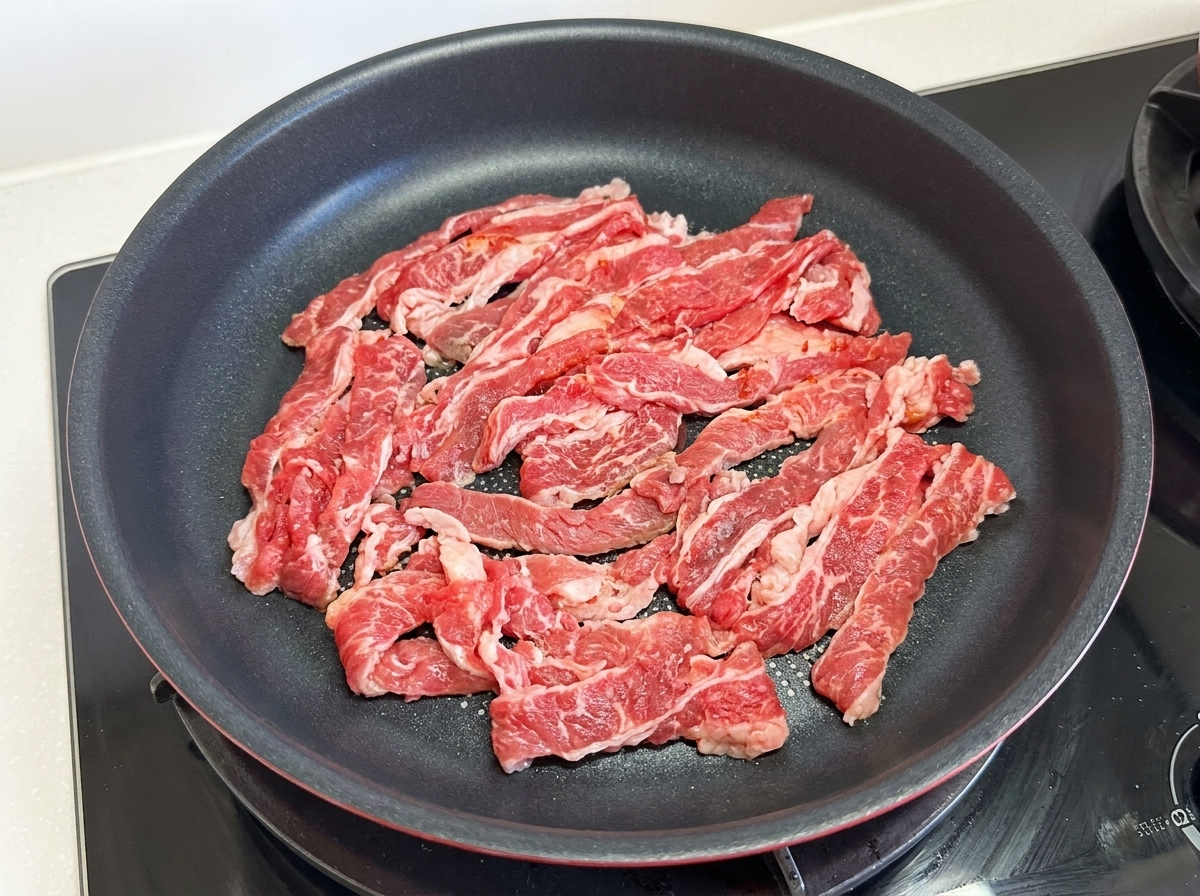 Fettine sottili di manzo grasso crudo distribuite sulla superficie di una padella antiaderente nera sul fornello.