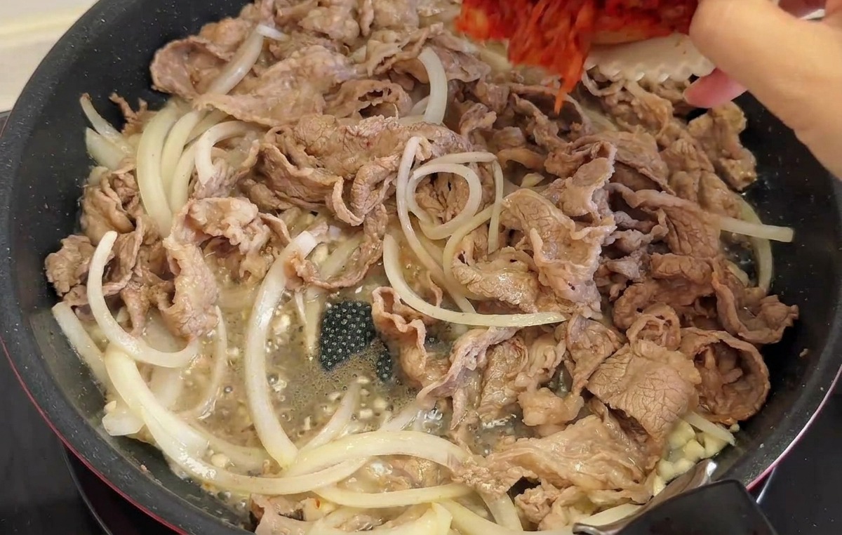 Una mano versa una ciotola bianca piena di kimchi rosso tritato e fette di peperoncino verde in una padella con manzo e cipolle.