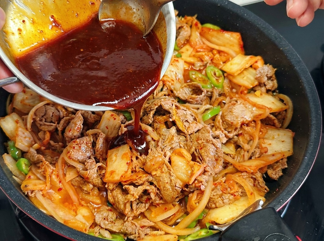 Una persona versa una salsa rosso scuro da una ciotolina di metallo in una padella contenente manzo, cipolle, kimchi e peperoncini.