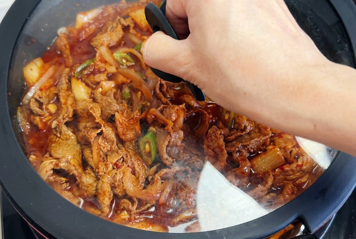 Una mano posiziona un coperchio di vetro con bordo nero sopra una padella piena di manzo al kimchi e peperoncini in salsa rossa.