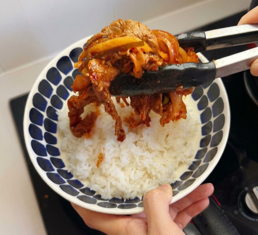 Pinze da cucina nere che sollevano una porzione di manzo cotto e kimchi rosso, adagiandolo su una ciotola di riso bianco.