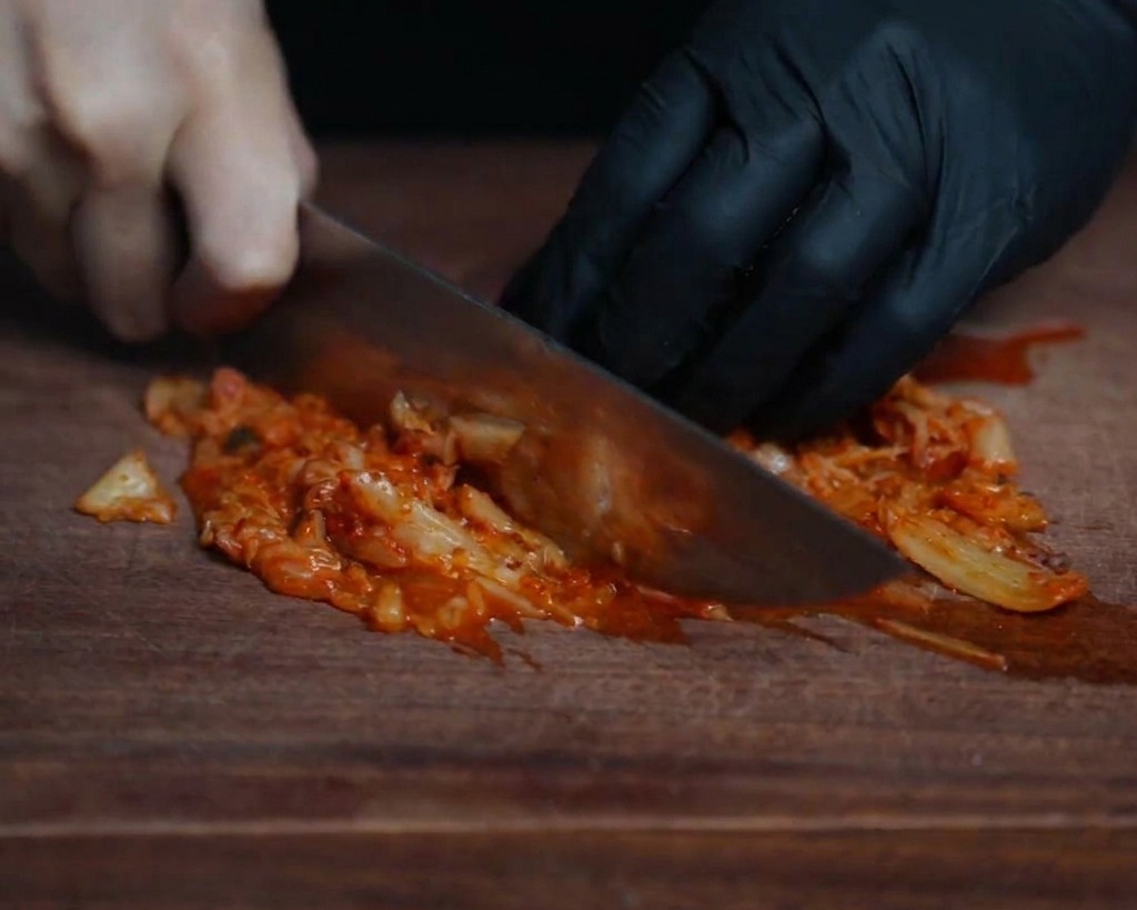 Primo piano di uno chef che taglia finemente il kimchi fermentato rosso brillante su un tagliere di legno scuro.