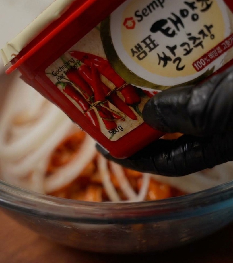 Una mano con un guanto nero tiene un contenitore di pasta di peperoncino sopra una ciotola per aggiungere una cucchiaiata di prodotto.
