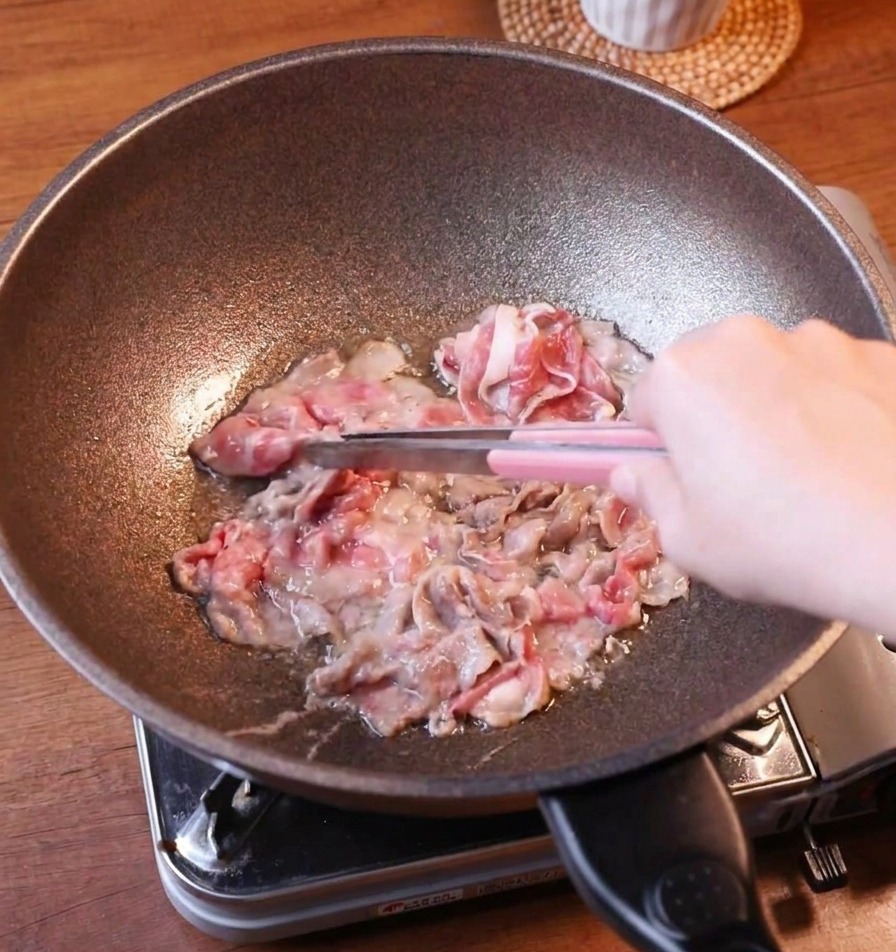 Fettine sottili di manzo dorato sollevate da una padella scura con pinze di metallo dal manico rosa.