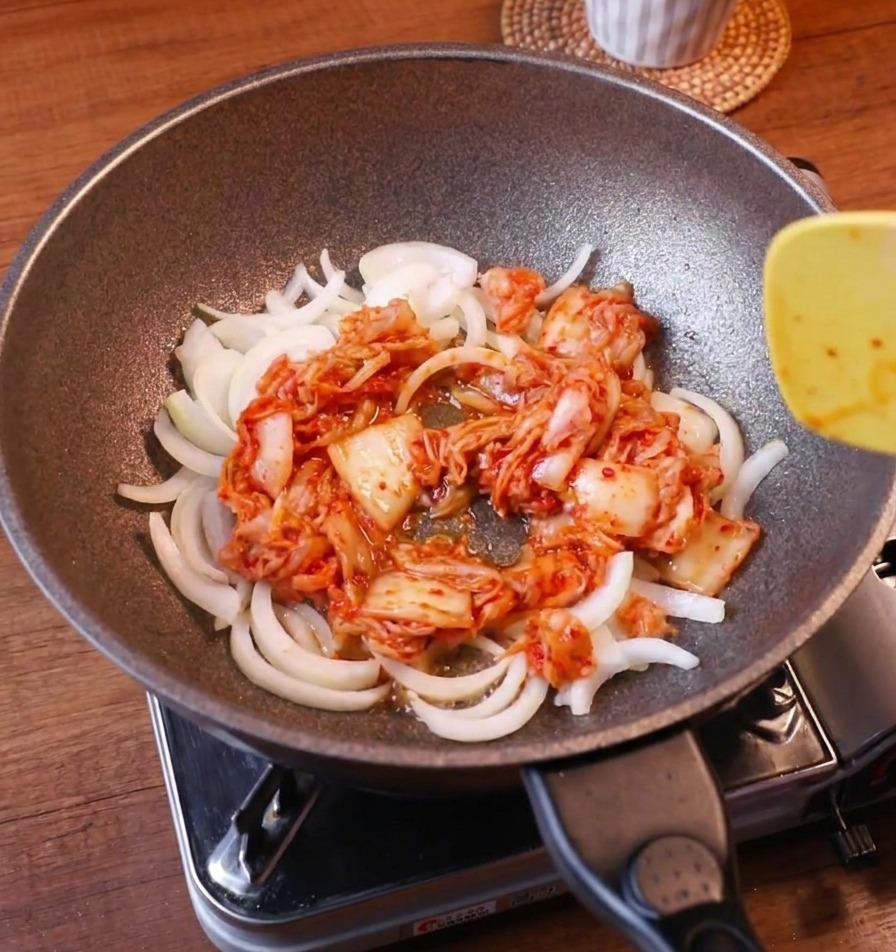 Cipolle bianche affettate e kimchi rosso acceso saltati insieme in padella con una spatola gialla.