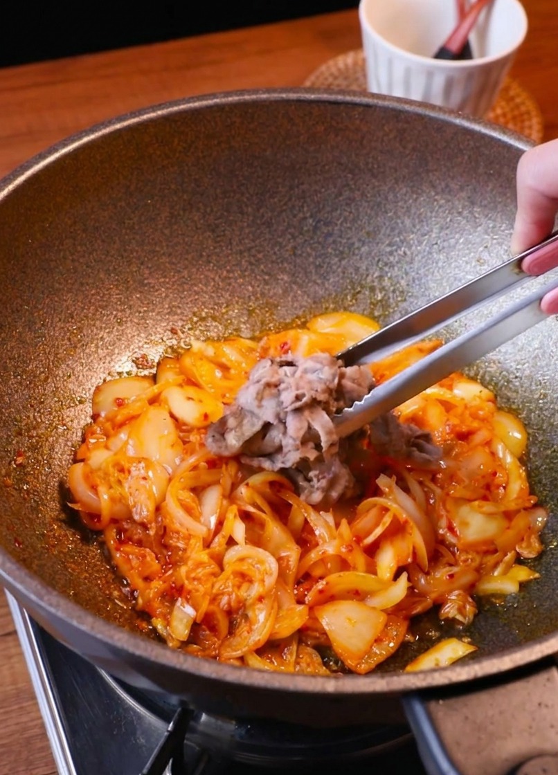 Delle pinze rimettono le fettine di manzo rosolate nella padella piena di cipolle e kimchi saltati.