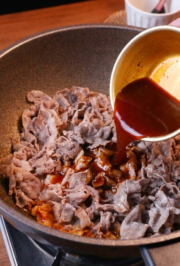Una gustosa salsa bruno-rossastra versata da una ciotola sopra la padella con manzo e kimchi.