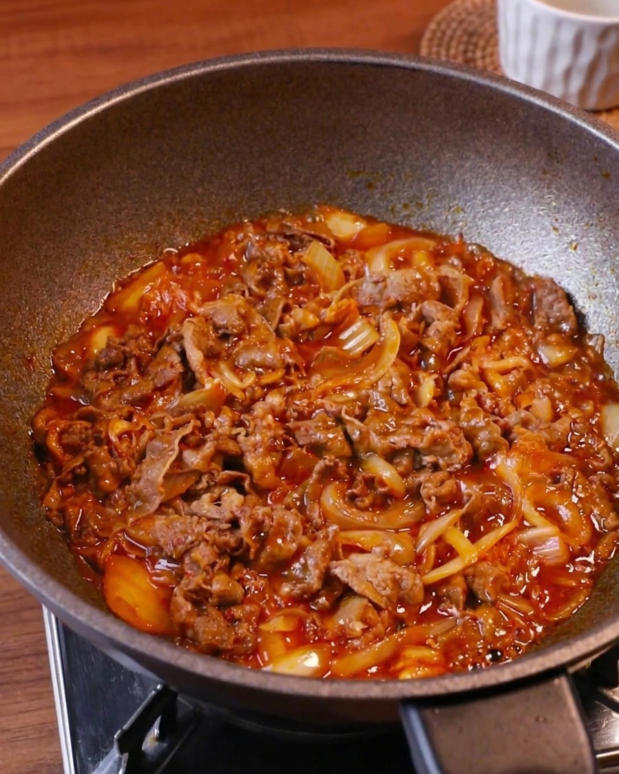 Teneri straccetti di manzo, kimchi e cipolle che sobbollono a fiamma alta in un wok con una ricca salsa rossa.