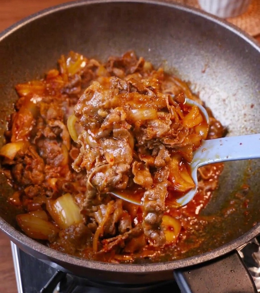 Un cucchiaio azzurro adagia una generosa porzione di manzo glassato al kimchi in una ciotola di riso bianco fumante.