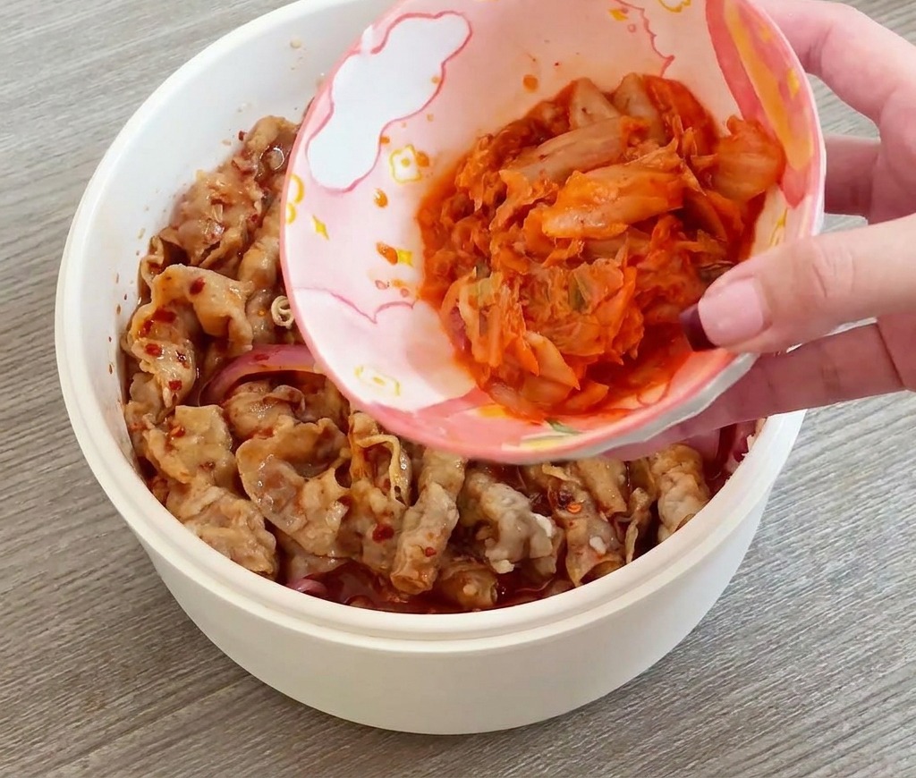 Una mano che versa una ciotolina di kimchi tagliato in una padella con manzo e cipolle.