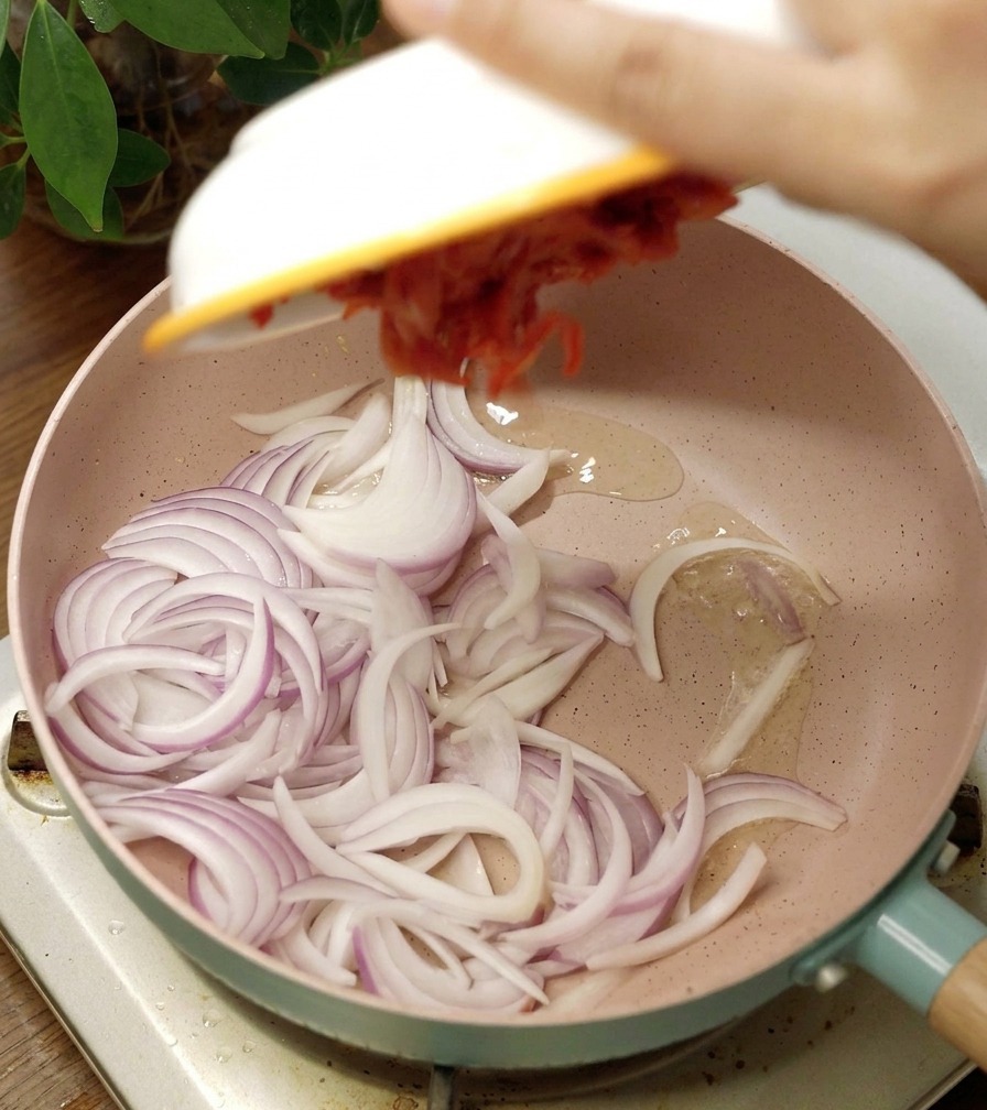 Una padella in ceramica rosa con cipolla rossa affettata e kimchi aggiunto da una ciotola bianca.