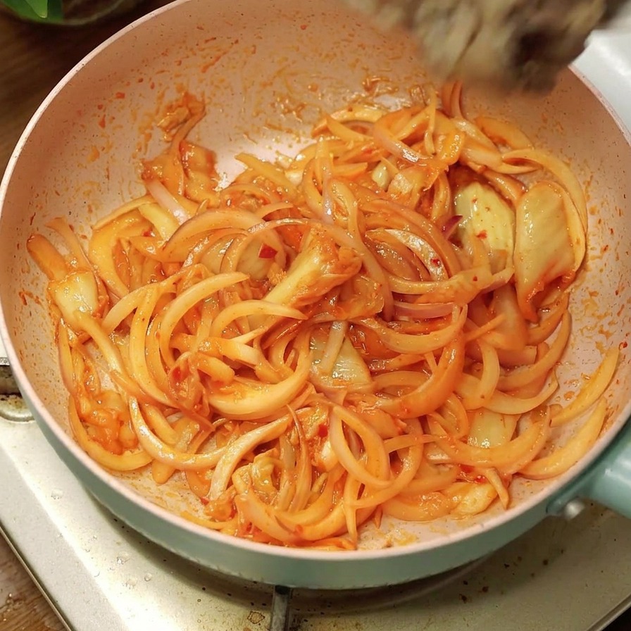 Fette di manzo cotte versate da una ciotola bianca in una padella con cipolle e kimchi.