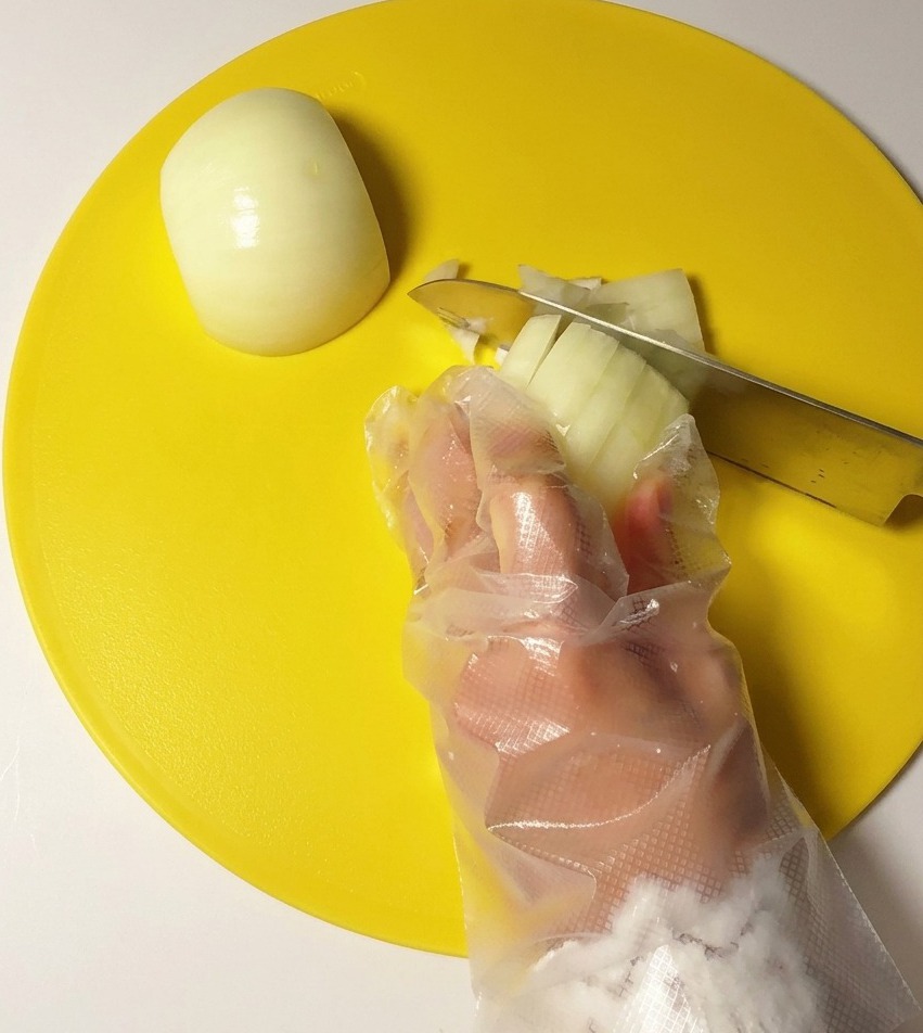 Una mano con un guanto di plastica trasparente taglia a cubetti una cipolla bianca con un coltello di acciaio inossidabile su un tagliere rotondo giallo.