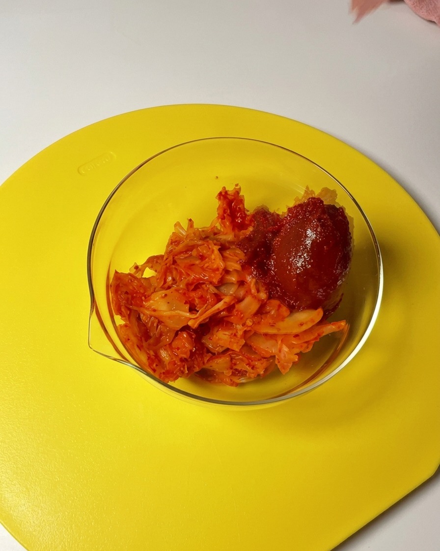 Un cucchiaio di gochujang rosso e peperoncino in polvere viene aggiunto a una ciotola di vetro piena di kimchi a pezzetti.