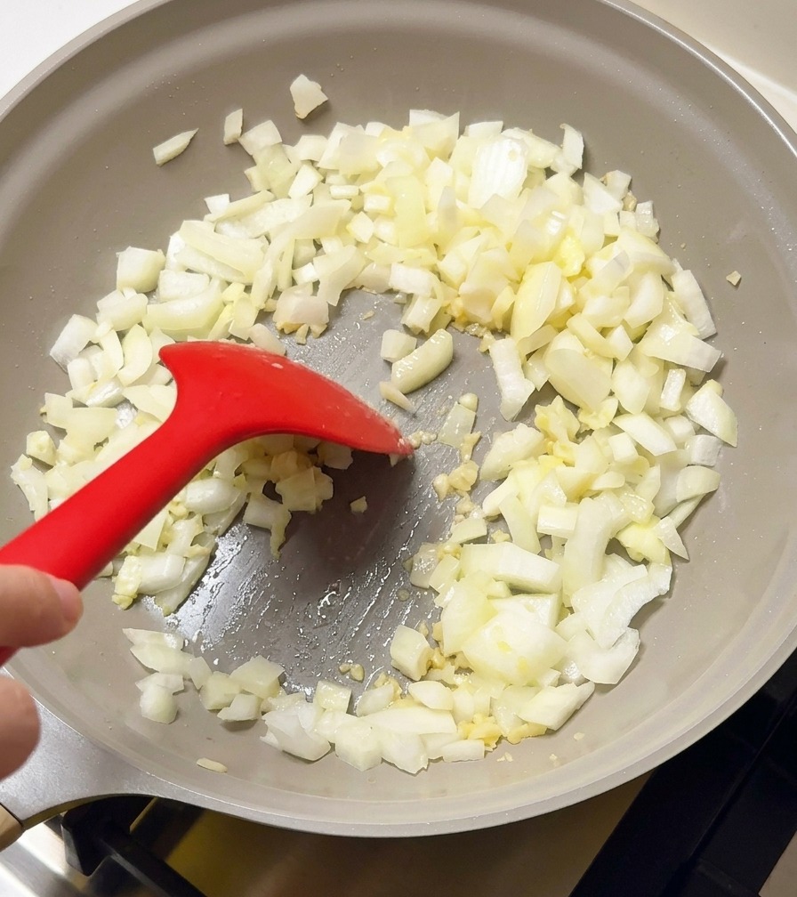 Cipolle bianche tritate e aglio vengono fatti saltare in una padella grigio chiaro con una spatola in silicone rossa.