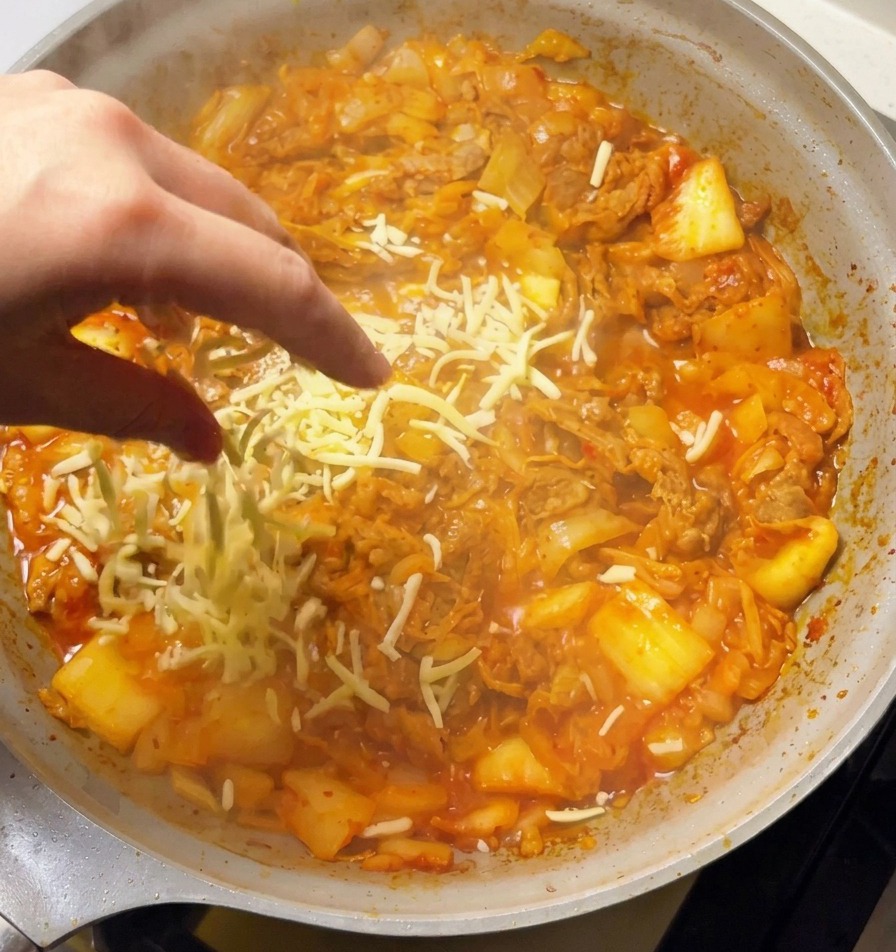 Una mano sparge abbondante mozzarella grattugiata su una padella bollente con il mix di manzo piccante e kimchi.
