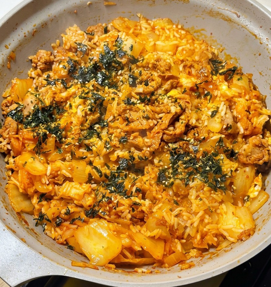 Una mano spolverizza fiocchi di alghe tostate verde scuro sopra il riso fritto fumante con filamenti di formaggio visibili.