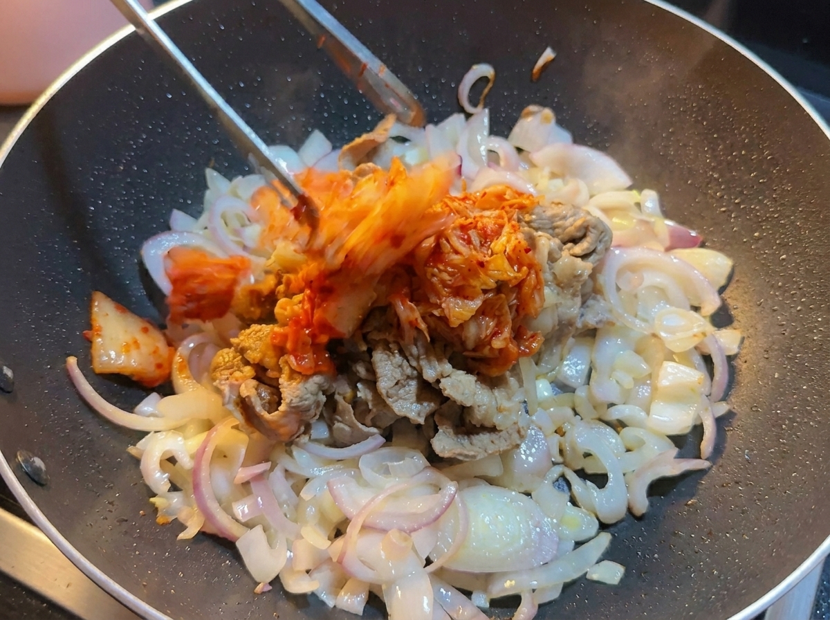 Kimchi di cavolo tritato aggiunto con le pinze in un wok contenente manzo e cipolle.