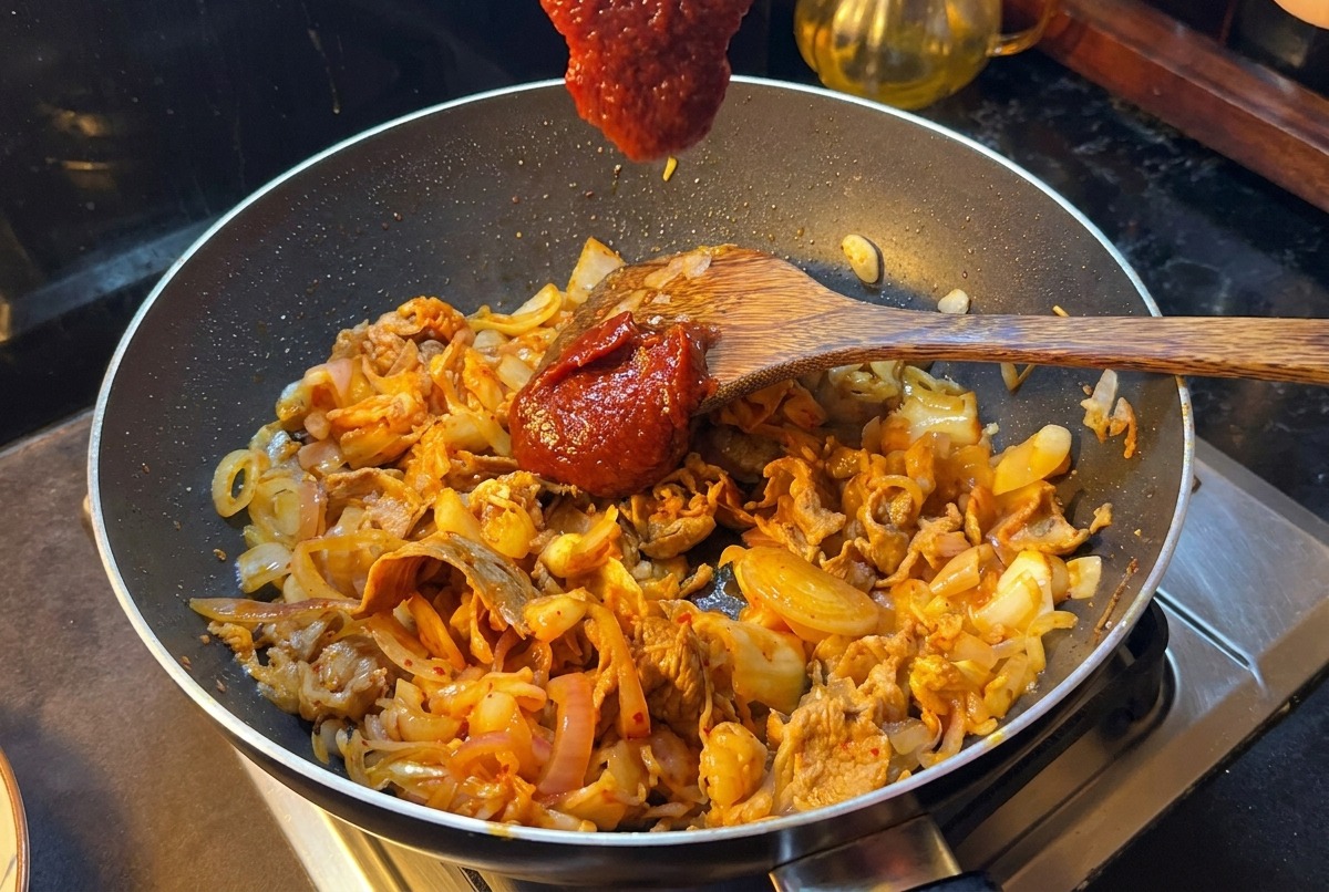 Una spatola di legno con una generosa cucchiaiata di pasta di peperoncino coreana sopra una padella di manzo, cipolle e kimchi.