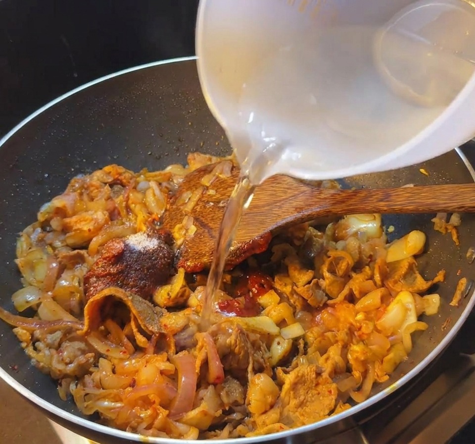 Acqua versata da una ciotolina in una padella nera che contiene manzo e kimchi in salsa rossa.