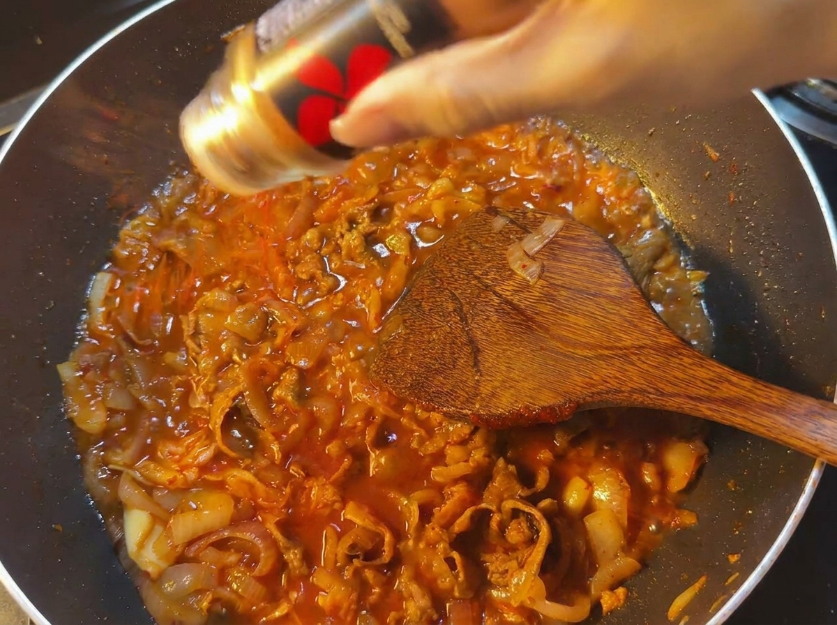 Peperoncino rosso in polvere versato da un contenitore nero in una padella di salsa rossa bollente con carne e cipolle.