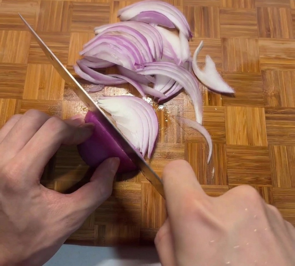 Una persona che affetta sottilmente una cipolla rossa su un tagliere di legno con un coltello da chef affilato.