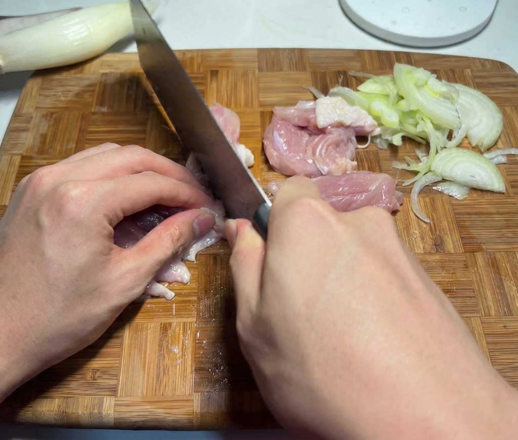 Uno chef che taglia cosce di pollo crudo a cubetti su un tagliere vicino a verdure affettate.