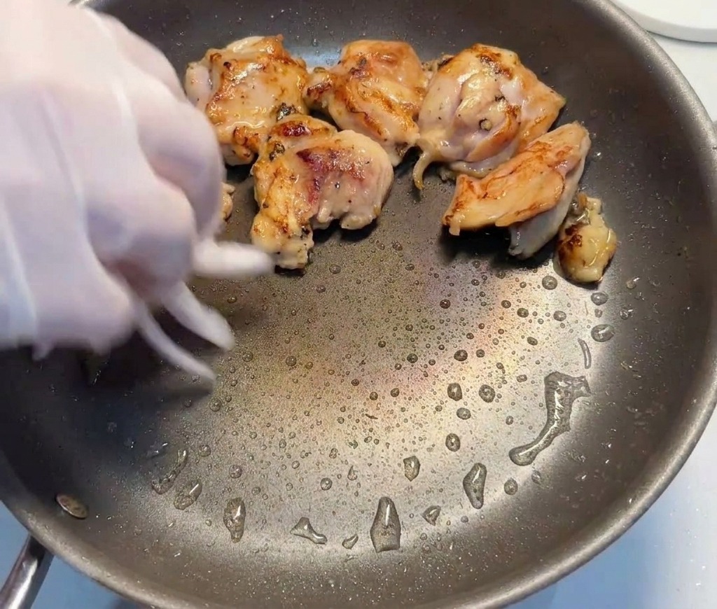 Una mano guantata che aggiunge fette di cipolla rossa in una padella con pezzi di pollo rosolati.