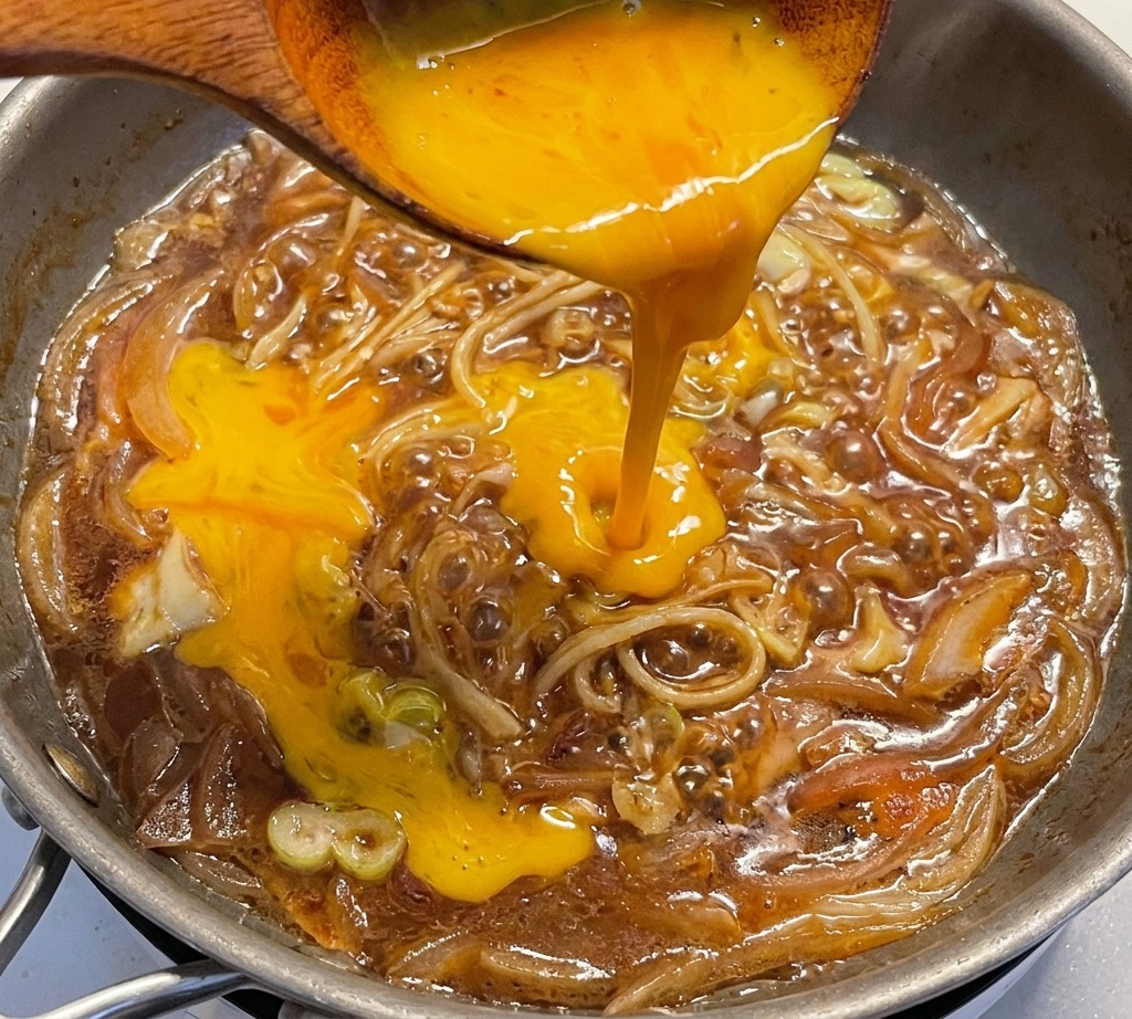 Uova sbattute versate da un mestolo di legno in una padella con cipolle e porri che sobbollono nella salsa.