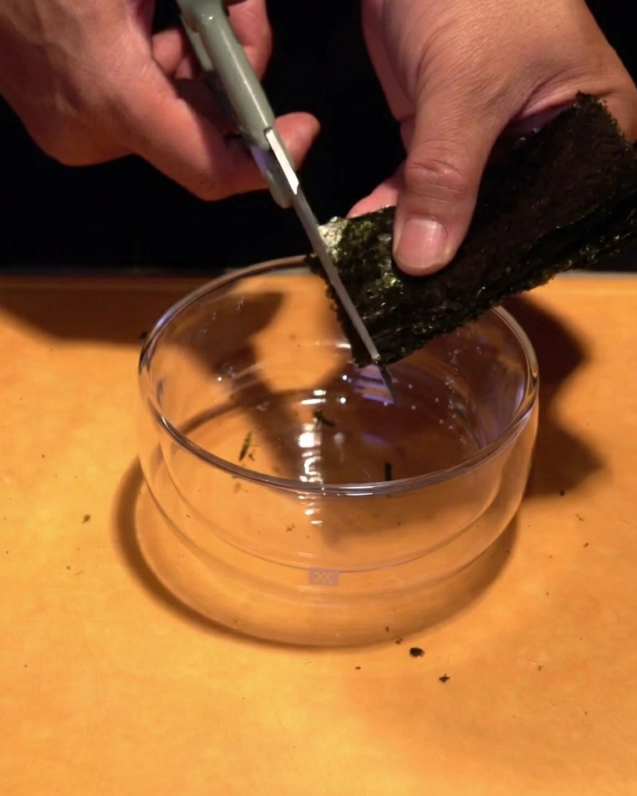 Mani che usano forbici da cucina per tagliare un foglio di alga verde scuro in strisce sottili sopra una ciotola di vetro.
