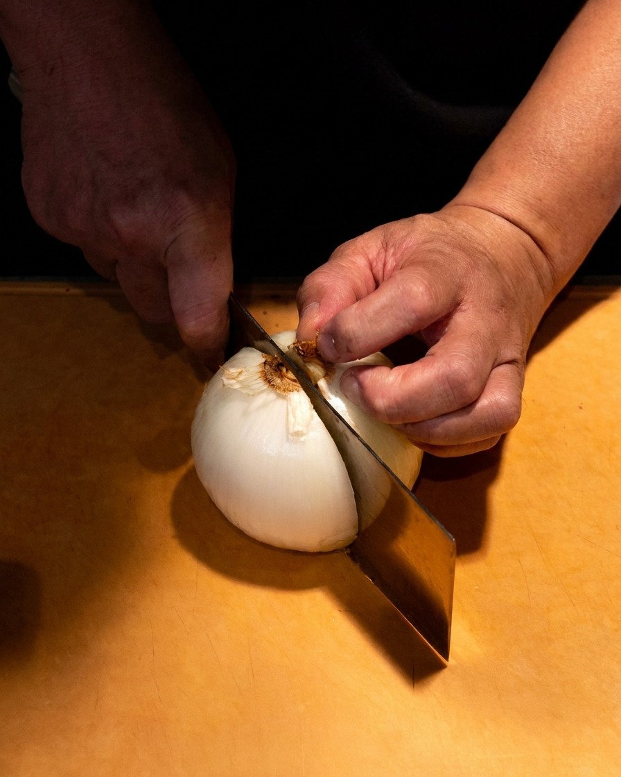 Primo piano di mani che affettano con cura una cipolla bianca in spicchi sottili su un tagliere di legno.