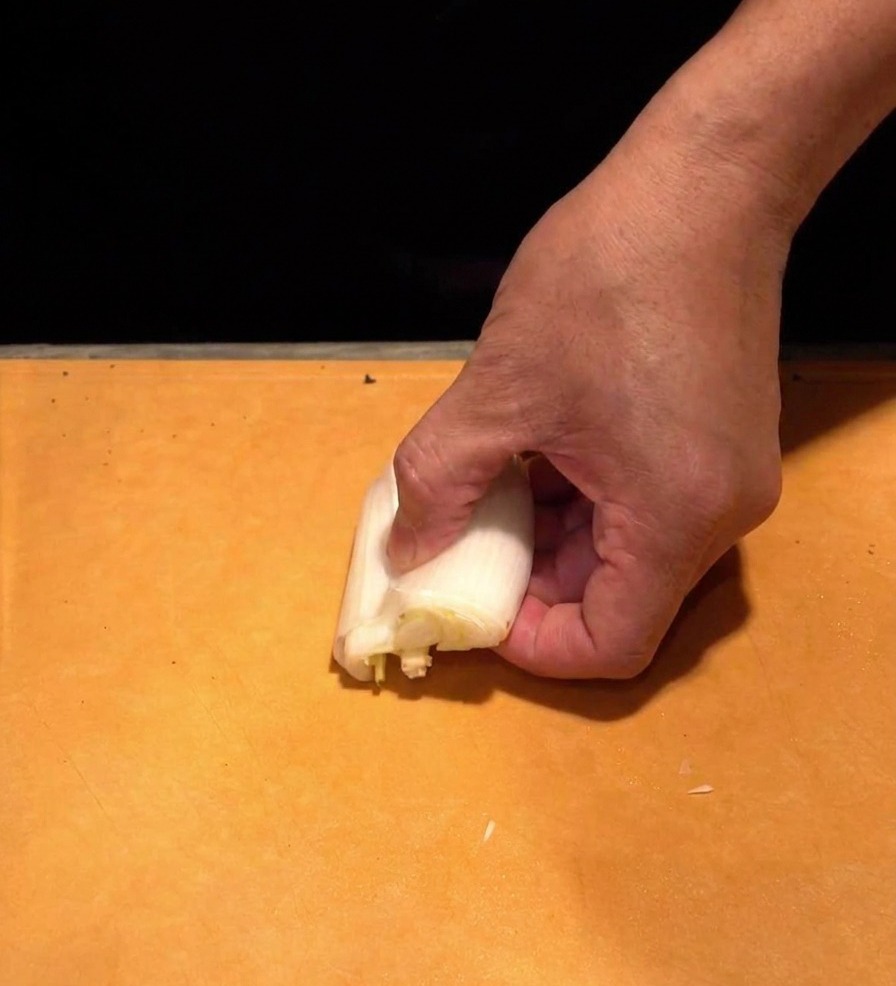 Una mano che tiene la parte bianca di un cipollotto naganegi su un tagliere, affettandolo in strisce bianche sottilissime.