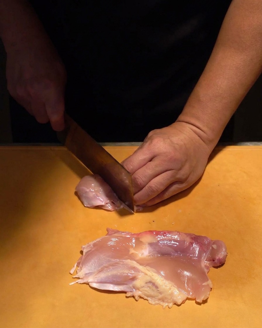 Carne cruda di sovracoscia di pollo tagliata a cubetti uniformi con un coltello affilato su un tagliere arancione.